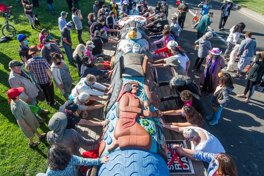A group of people reach out and touch a pole, which is lying sideways along a truck bed, painted in bright colors with a figure of a kneeling woman