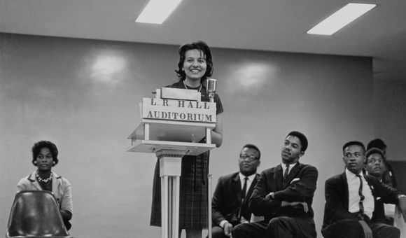 A black-and-white image of Diane Nash at the podium with men and a woman sitting on either side of her in chairs. Nash stands in the center of the image behind the podium labeled [L.R. HALL/ AUDITORIUM] speaking into the microphone.