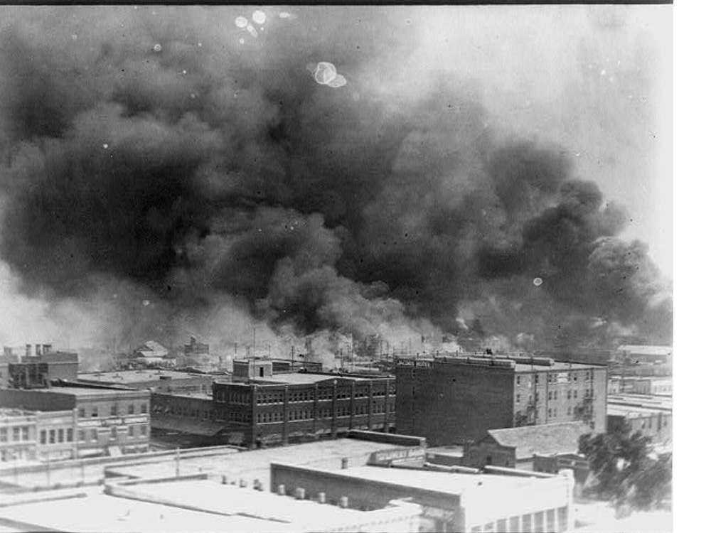 A black and white photo of buildings, from an aerial perspective, with thick plumes of black smoke rising above and obscuring the sky