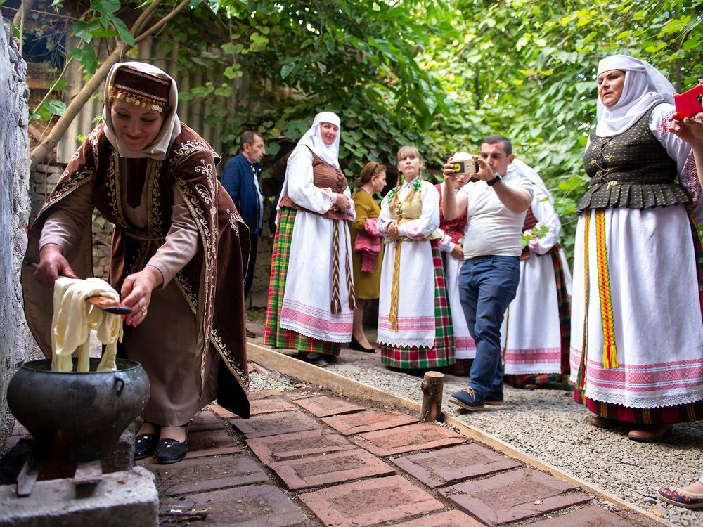 A woman wearing traditional Armenian dress bends down to tend to an outdoor stove while preparing a meal.