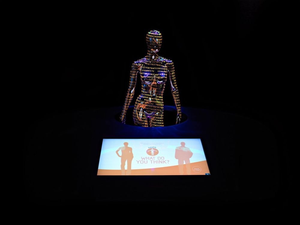 A wide shot of a museum exhibit, featuring a dark, faceless, naked female mannequin covered in orange genetic code in the background with a monitor in the foreground showing an orange and white graphic of two people with "What do you think?" between them.