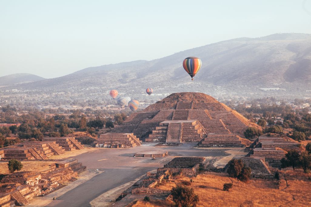 In an aerial view of an ancient city, a hot air balloon floats above an enormous stone temple with steps leading up to its peak