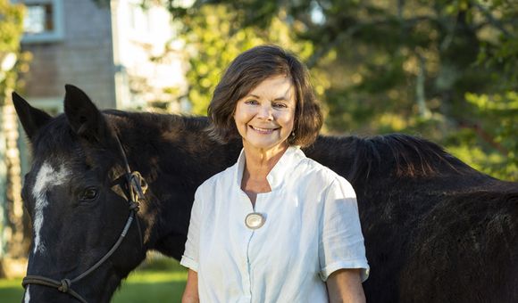Image shows a woman wearing a white shirt standing with and holding the reins of a horse