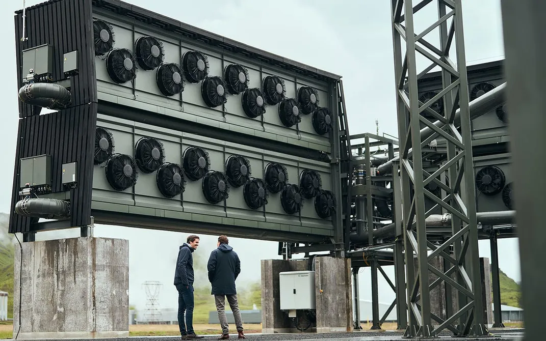 Two men stand in front of tall wall of fans at the Orca carbon capture facility in Iceland