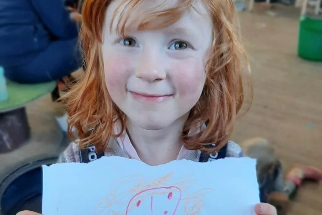 A pale, red-headed girl with bangs smiles and holds a piece of paper in front of herself, with what looks like a drawing of a person's smiling face and orange curly hair