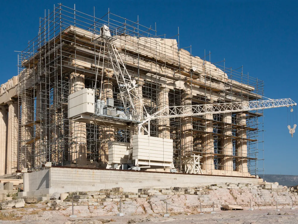 A view of the side of the Parthenon, on a sunny day with clear blue sky behind; the facade is covered in scaffolding and construction equipment