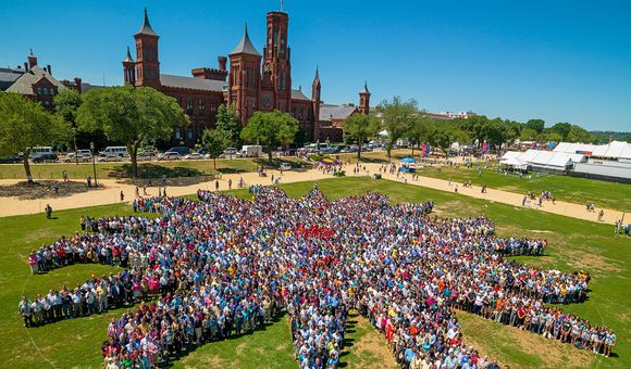 Thousands of people assemble on the green grass of the National Mall to form a sunburst shape, the Smithsonian logo. The red-brick Smithsonian Castle rises in the background.
