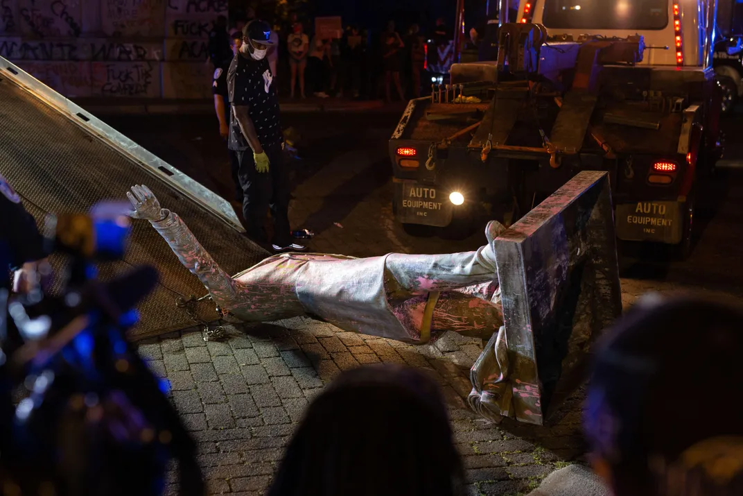 Surrounded by flashing lights at night, a paint-splatted statue of a man is lying on its side and being towed onto a truck