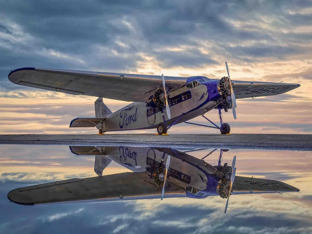 A vintage silver Ford Tri-Motor aircraft parked on a wet tarmac under a dramatic, cloudy sky at dusk. The plane features corrugated metal skin, three engines, and the "Ford" script logo in blue on the fuselage, all reflected in the water on the ground.