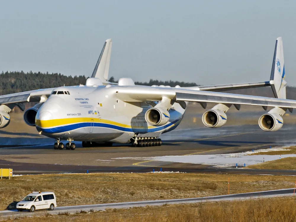 A photo of the world's largest plane, the Antonov AN-225. It is sitting in an airfield. The plane is mostly white and has yellow and blue stripes.