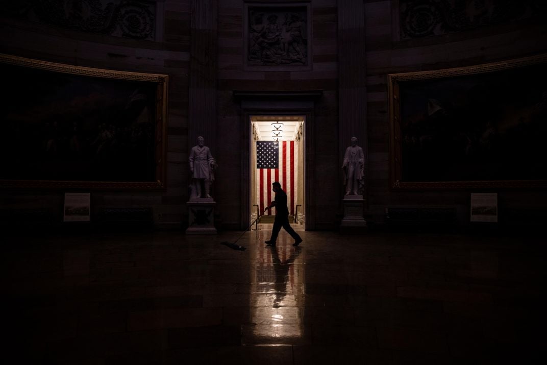 In a dramatic shot, a worker walks across a strip of light from a door behind him as he cleans the floor; the lights appear to be off, and framed by the dark, the worker's shadow and an American flag are starkly visible in the center of the frame 