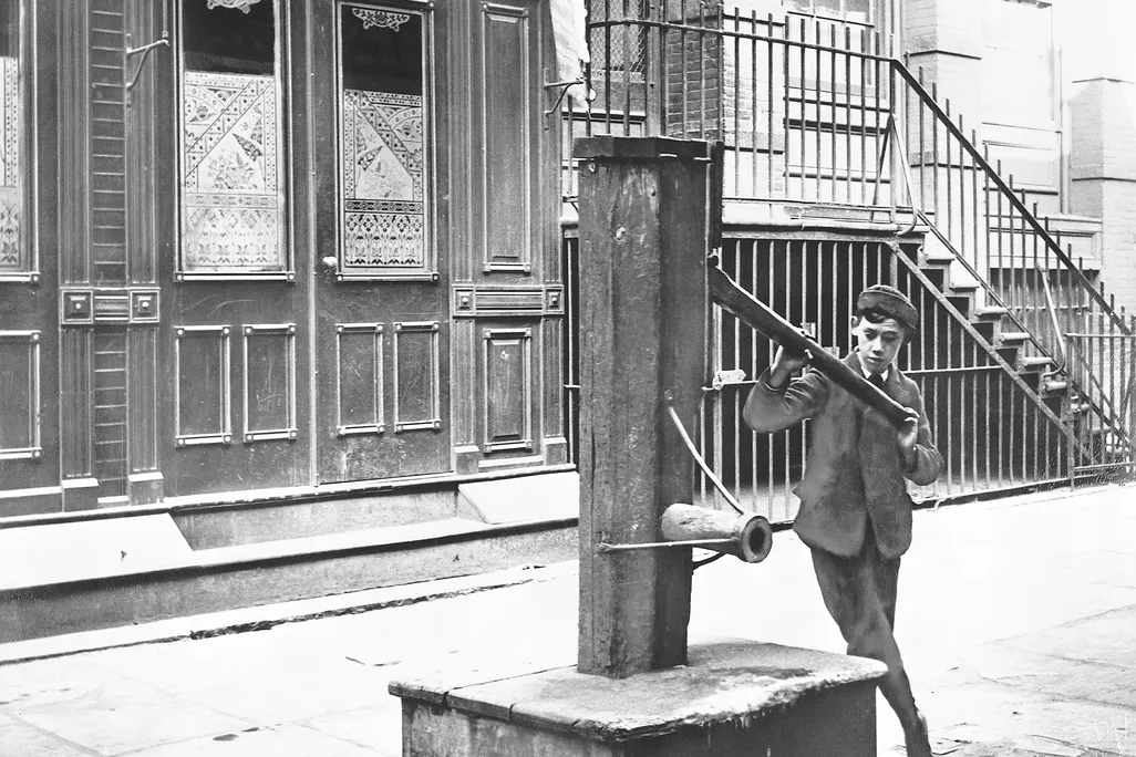 An old photo of a boy holding the handle of water pump on a street in New York City
