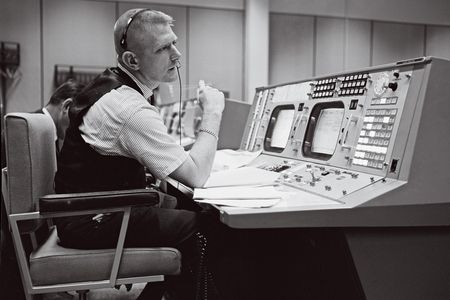 A black-and-white image of a white man in his early 40s sitting at a work console with two display screens and dozens of buttons. The man is wearing a short-sleeve shirt covered by a black vest.