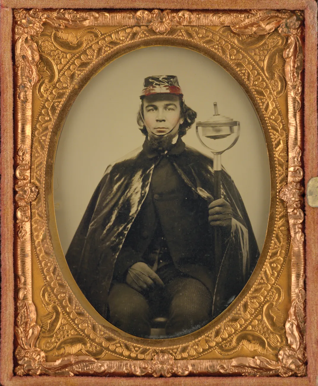 a tintype portait of a man in oilcloth cape, a soldier’s cap holding an oil torch
