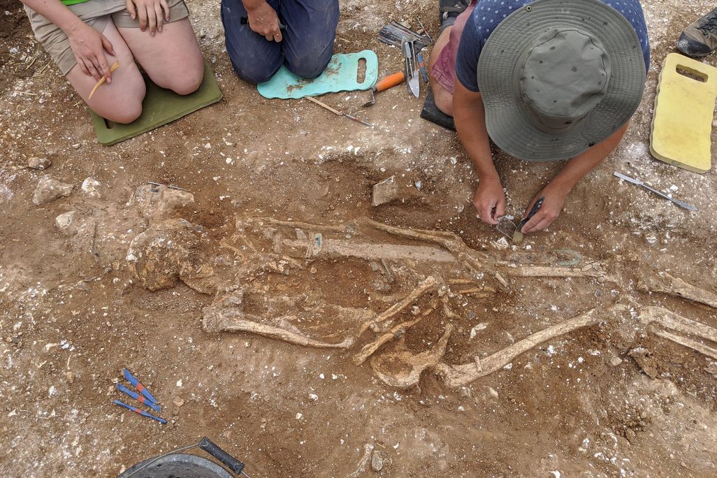 Three people kneel and bend to look at the grave; one person wearing a hat appears to be delicately touching or removing part of the skeleton