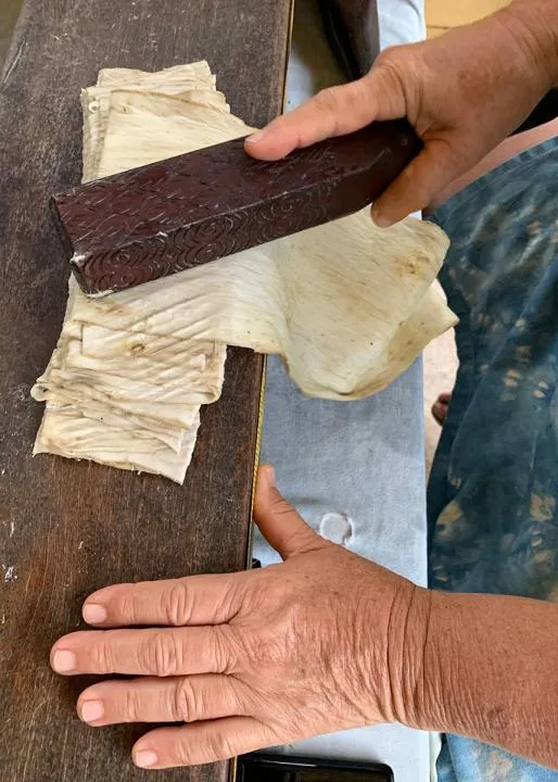 Close-up on a pair of hands, one holding a four-sided dark wooden beater against a folded sheet of pale bark cloth.