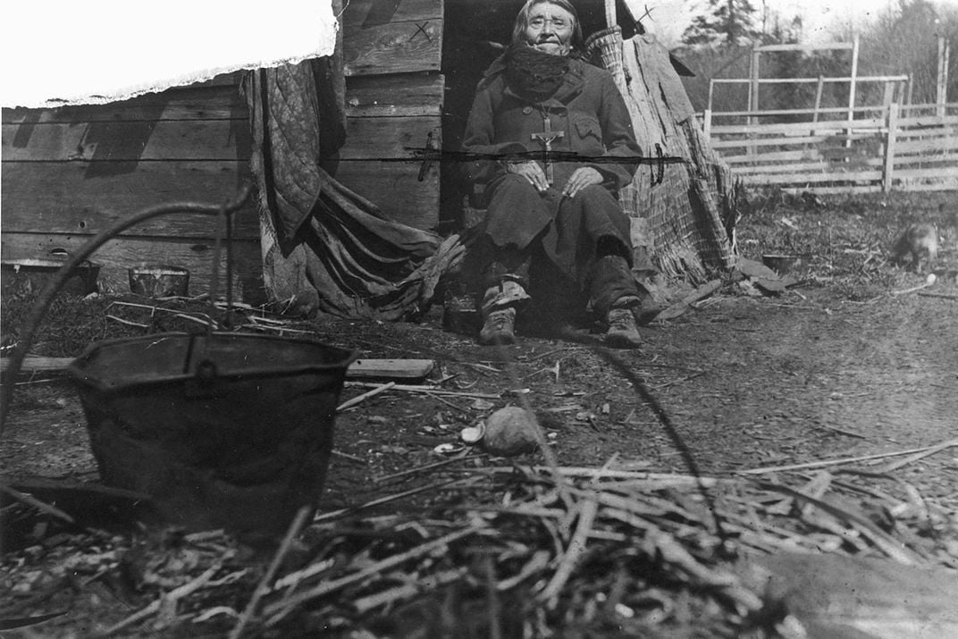A man sits in the doorway of a wooden shack. In the foreground is an outdoor fire pit and black pot. Black-and-white archival photo.