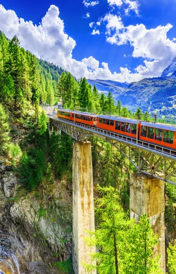 Train Journeys - a sunlit train crossing a bridge over a chasm on a forested mountain with a waterfall in the background.
