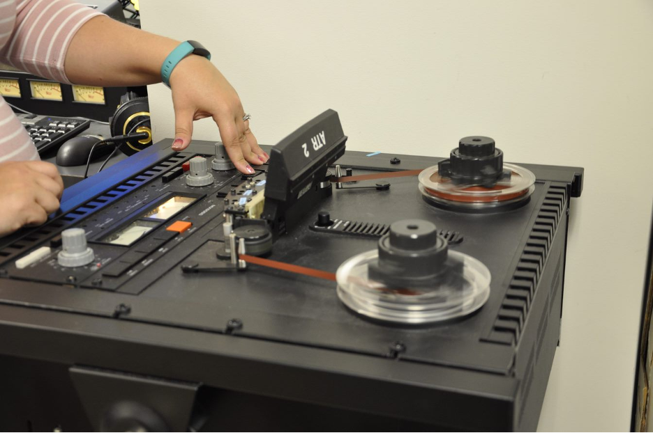 A black piece of equipment has two plastic hubs of magnetic audio tape on top, with two hands on the left side of the image controlling the equipment using multiple gray knobs.