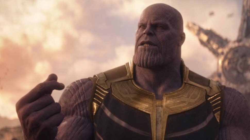 Thanos holds his right hand up and crosses two fingers to form the beginning of a snap. Behind him is a hazy sky.