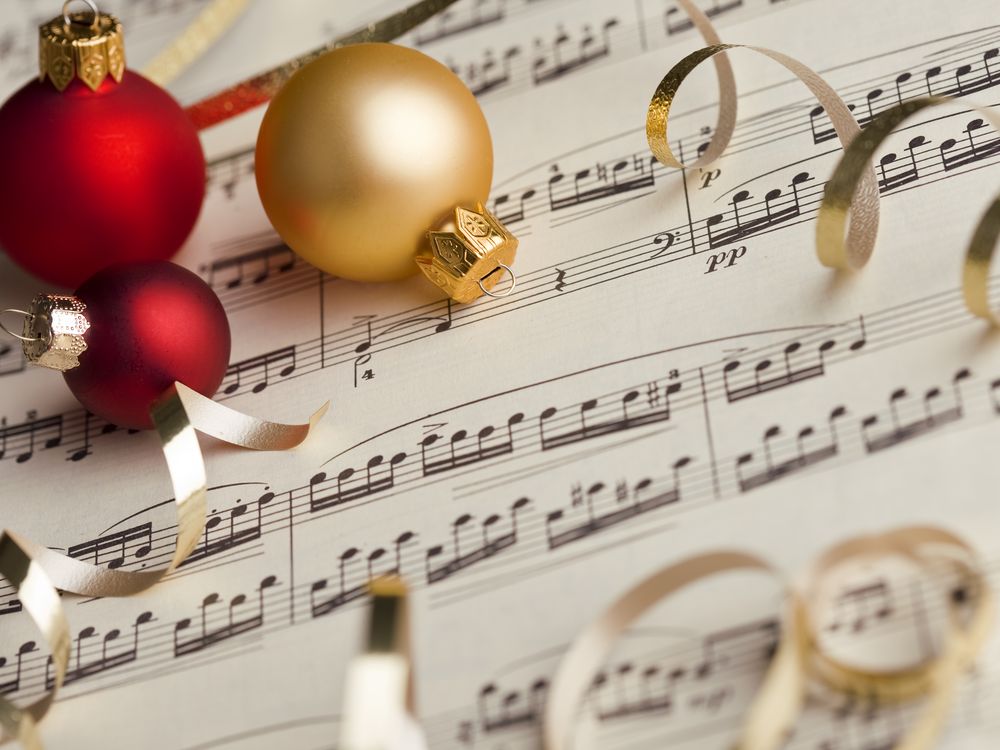 Red and gold Christmas ornaments rest on top of open sheet music, surrounded by curled gold ribbon, creating a festive holiday scene