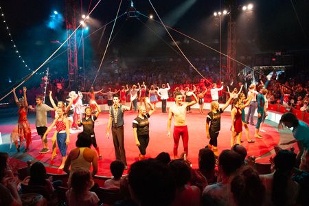 Performers wave, forming a ring around a red, circular stage, with a packed audience and rigging hanging above.