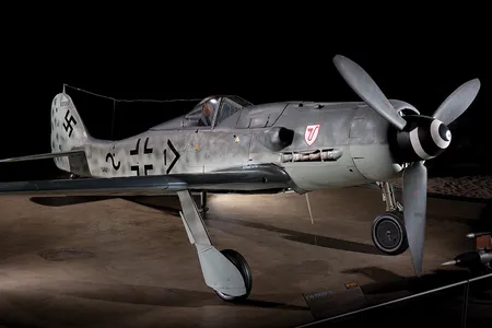 A German fighter aircraft is painted dull grey, with some black markings including a Nazi swastika. A large gun in mounted near the nose of the plane, which has a single large propeller.