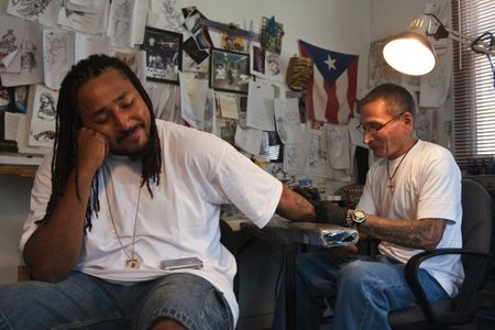 Q Monts sits patiently at Nu Flava Ink while tattoo artist Charles “Coco” Bayron puts the finishing touches on Q’s tattoo reading “Time is Money, Money is Time.”