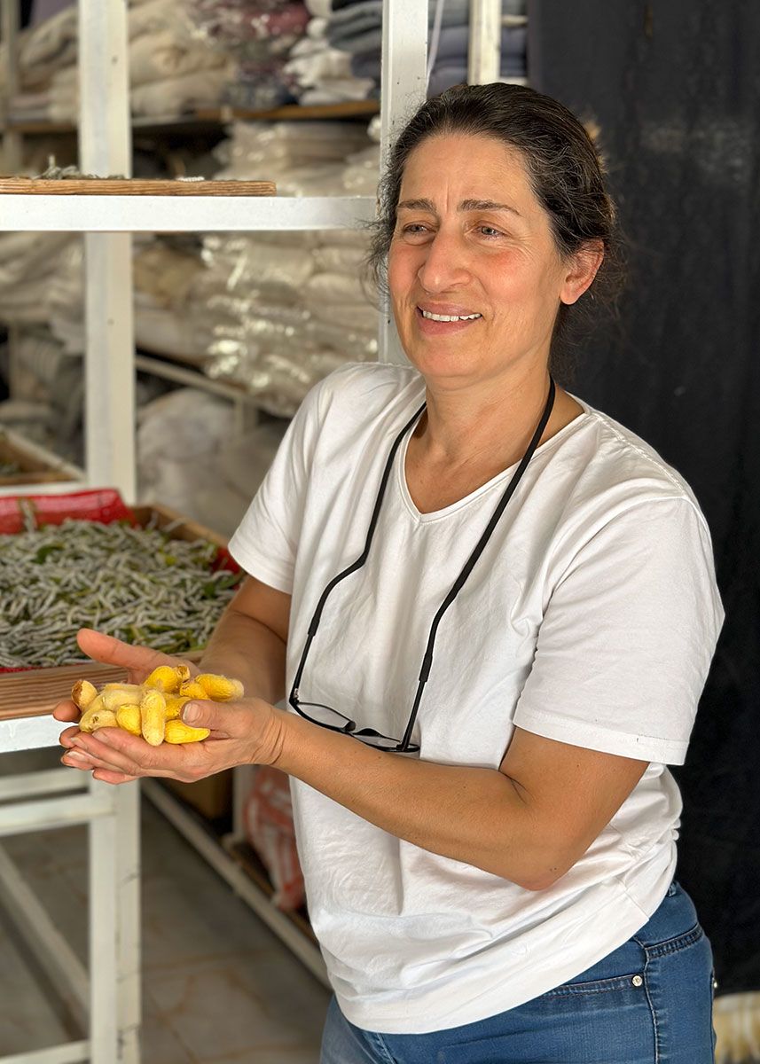 A woman holds up a handful of yellow silkworm cocoons, with trays of silkworms behind her.