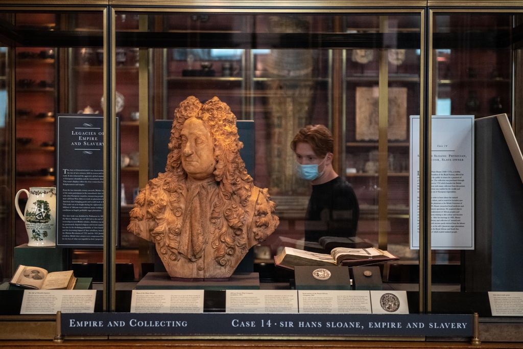A glass case with the newly positioned bust of Sloane, which is a terracotta color, next to a small sign that has paragraphs of text explaining how he profited from enslavement. Behind, a visitor walks by wearing a mask.