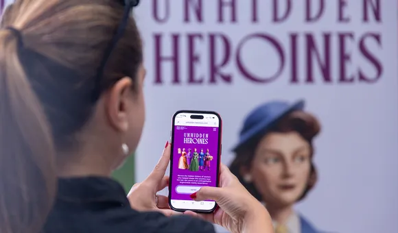 View from the back as a woman pulls up a webpage on her phone. In the background a poster reading “Unhidden Heroines” is visible.