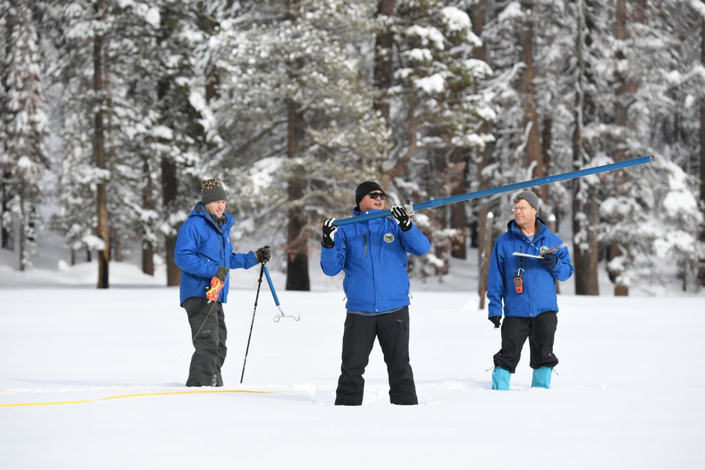 Three men in blue coats standing in the snow with equipment