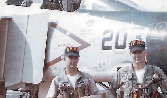 Two U.S. Navy men in uniform--a pilot and an aviator — stand in front of the cockpit of an F-4B Phantom II military jet aircraft.