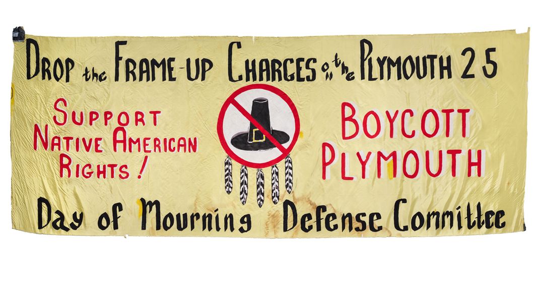 Large handmade banner that shows a Pilgrim hat framed in a prohibition sign, decorated with feathers. The banner reads: “Drop the Frame-Up charges on the Plymouth 25. Support Native American Rights! Boycott Plymouth. Day of Mourning Defense Committee.”
