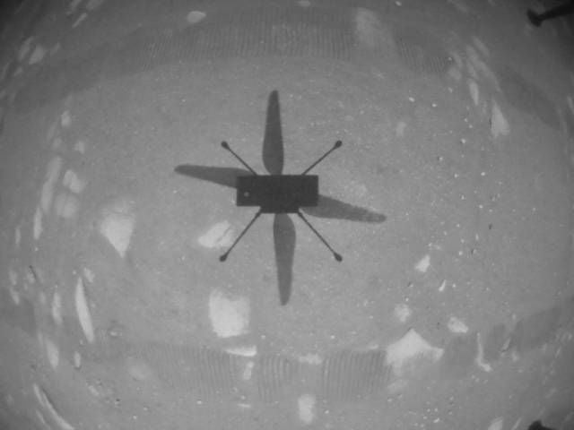 An image taken by the Mars Helicopter Ingenuity during it's first test flight. The image is black and white and shows the helicopter's shadow on the Martian surface.