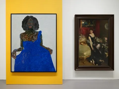 A painting of a Black woman in a blue dress is hung against a yellow backdrop, next to it is a portrait of a white woman sitting on a green couch.
