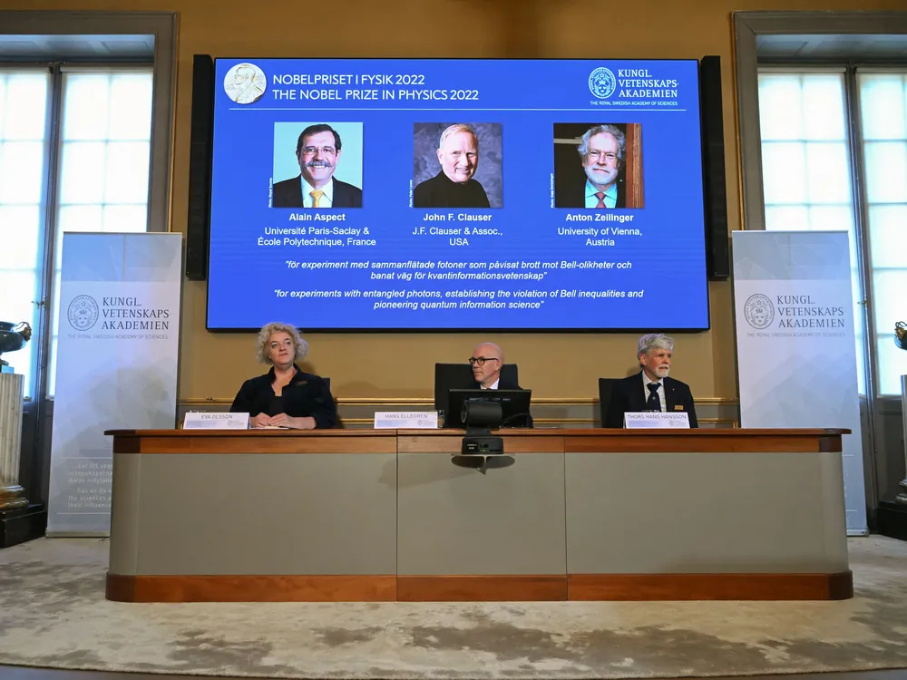 Members of the Nobel Committee for Physics seated at a table, with a screen displaying pictures of the winners of the prize behind them.