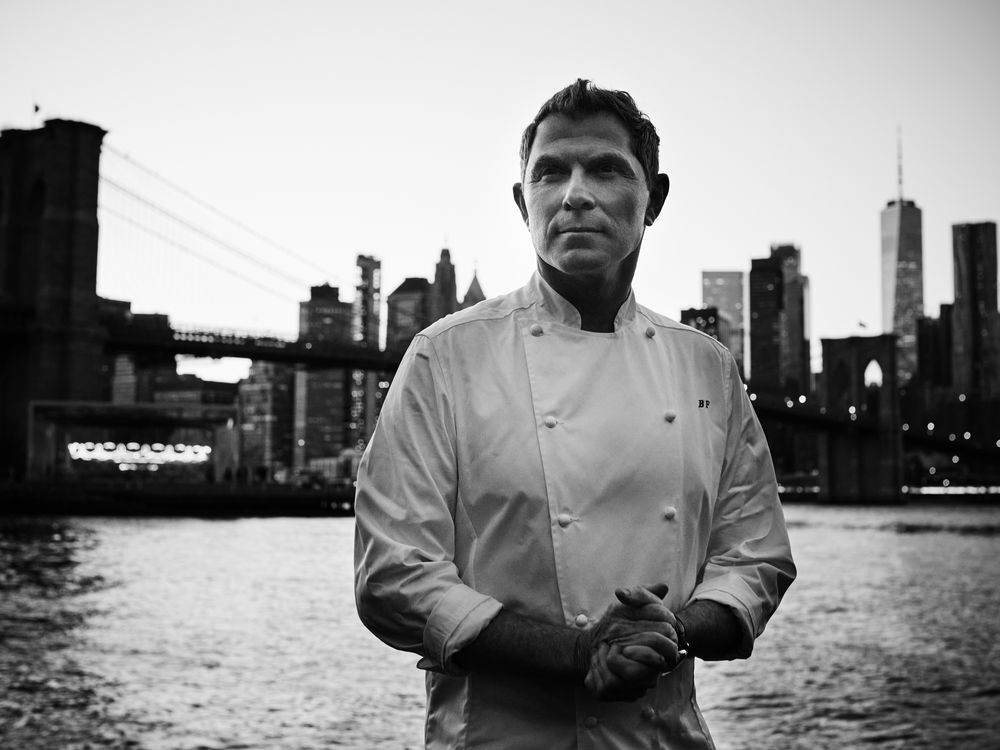 A black and white photograph of Bobby Flay in his signature white chef’s coat, with his initials, BF, embroidered on the front.