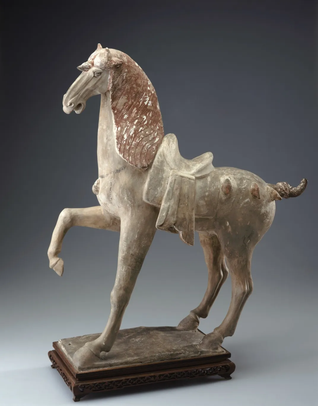 A stoneware sculpture of a horse with one hoof in the air and decorative tassels on the body and one on the forehead.