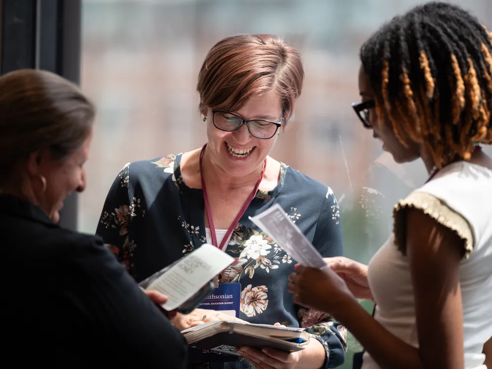 Three women stand in a circle laughing and discussing printed handouts