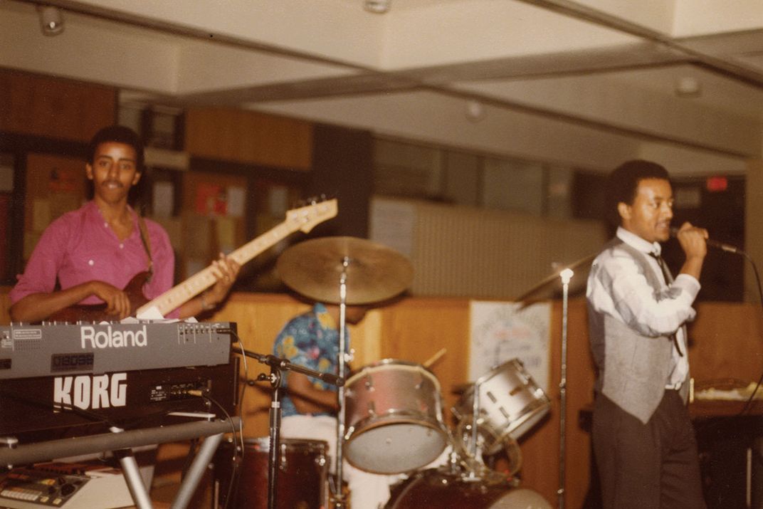 Three band members performing on electric bass, drums, and vocals. Old color film photo.