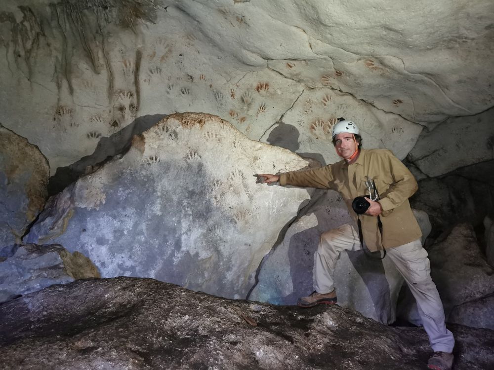 A man wearing boots and a white helmet stands in front of a large rock and points to handprints, which are reddish or white against black shadows and cover the sloping wall of the cave