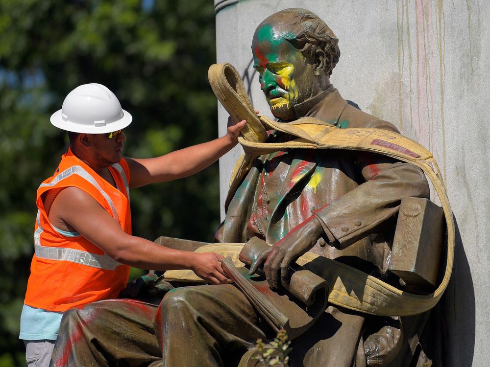 A man in a bright orange construction vest and white hat wraps a thick rope around a paint-splattered statue, of a bearded man wearing a suit and sitting in a chair