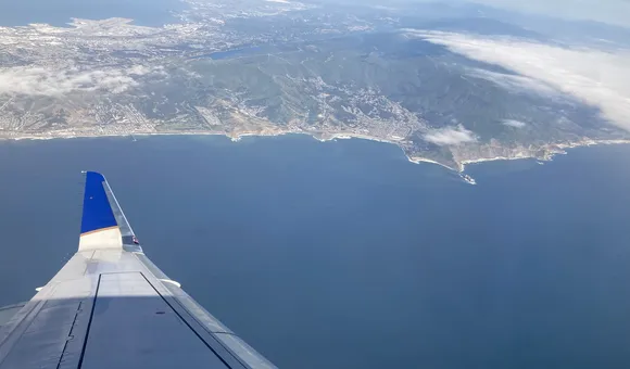 View from a plane’s window showing the wing as the plane flies over the ocean and shoreline.