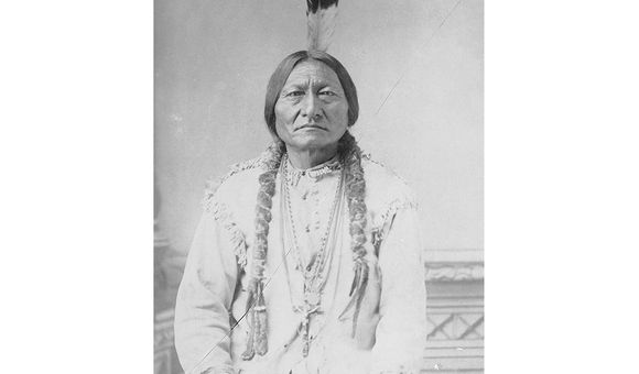 Historic black and white photograph of Sitting Bull sitting down with a feather in his hair and holding a pipe.