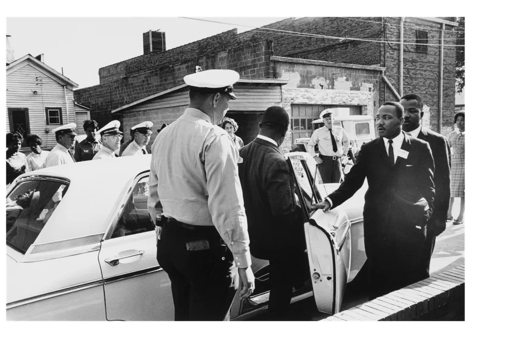 A black and white photograph of policeman in foreground, one man stepping into a squad car and MLK with an arm outstretched moving toward the car