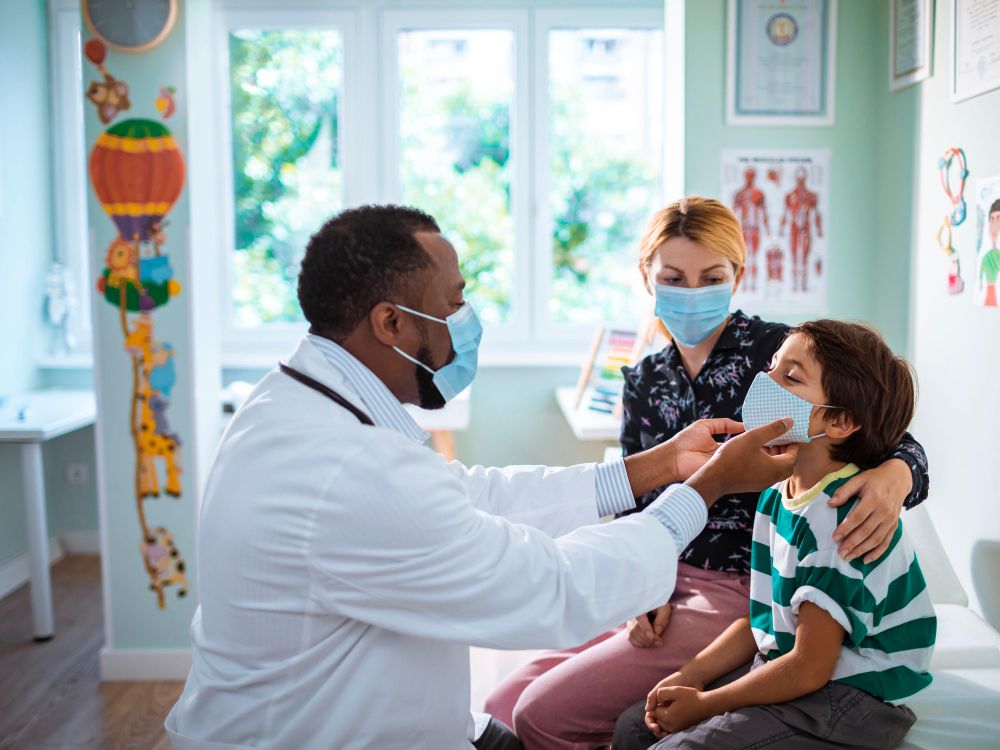 A child sits next to a woman with blond hair while a pediatrician looks at his face. All three are wearing masks.