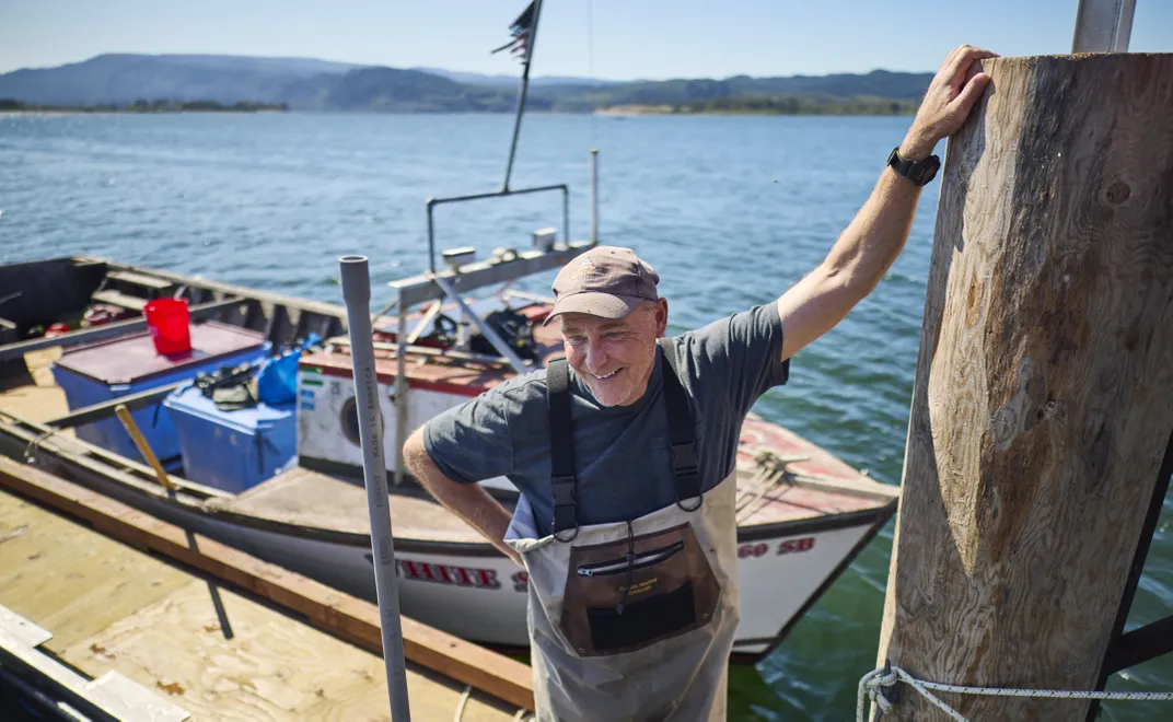 a man stands on a dock, leaning into a piling, with a small boat in the background