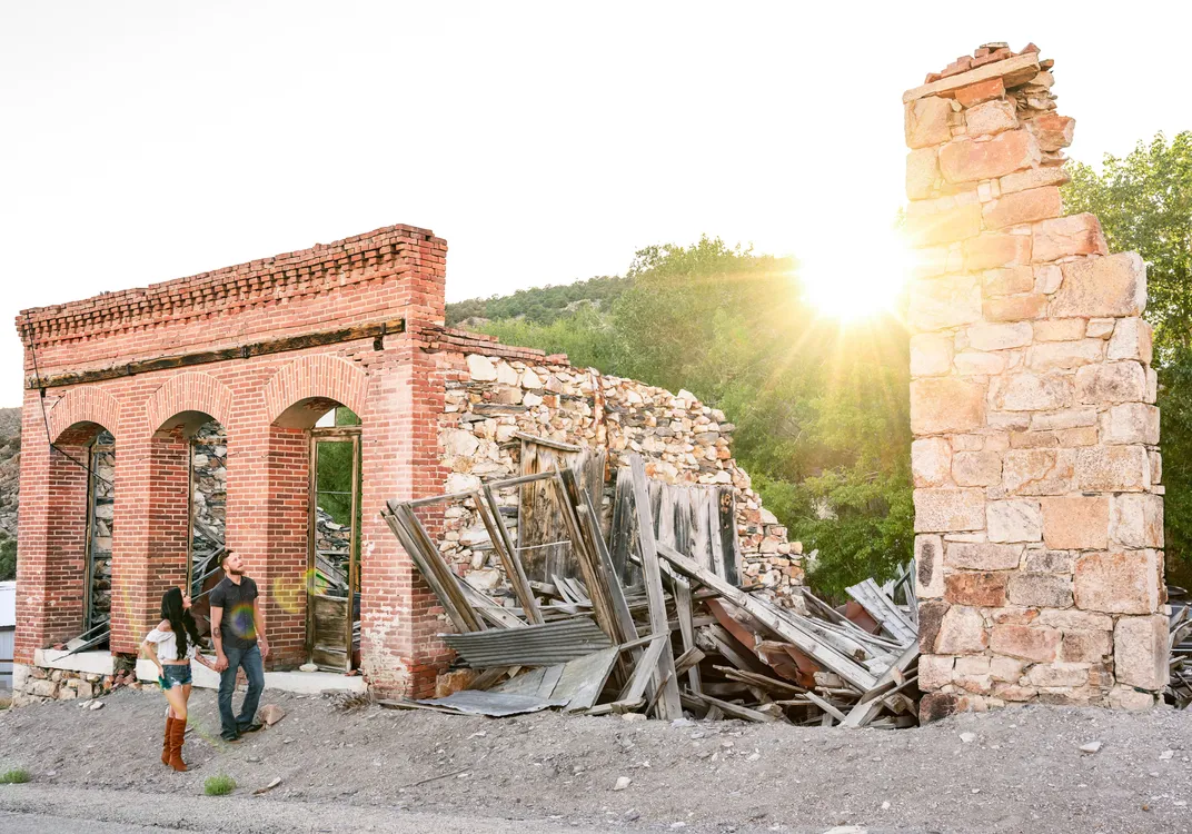 Partially collapsed brick and stone building with three arched doorways at sunset; two people stand nearby, one in a hat and jeans, the other in shorts and boots.