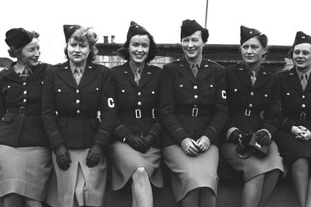 Six women sitting closely together posing for the camera. They are wearing uniforms with light skirts and dark jackets and an armband with the letter C on it.